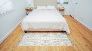 Ein ordentlich bezogenes Bett mit beige-farbenem Kopfteil, zwei Nachttische mit Lampen und ein Streifenmuster-Teppich auf Holzdielen.