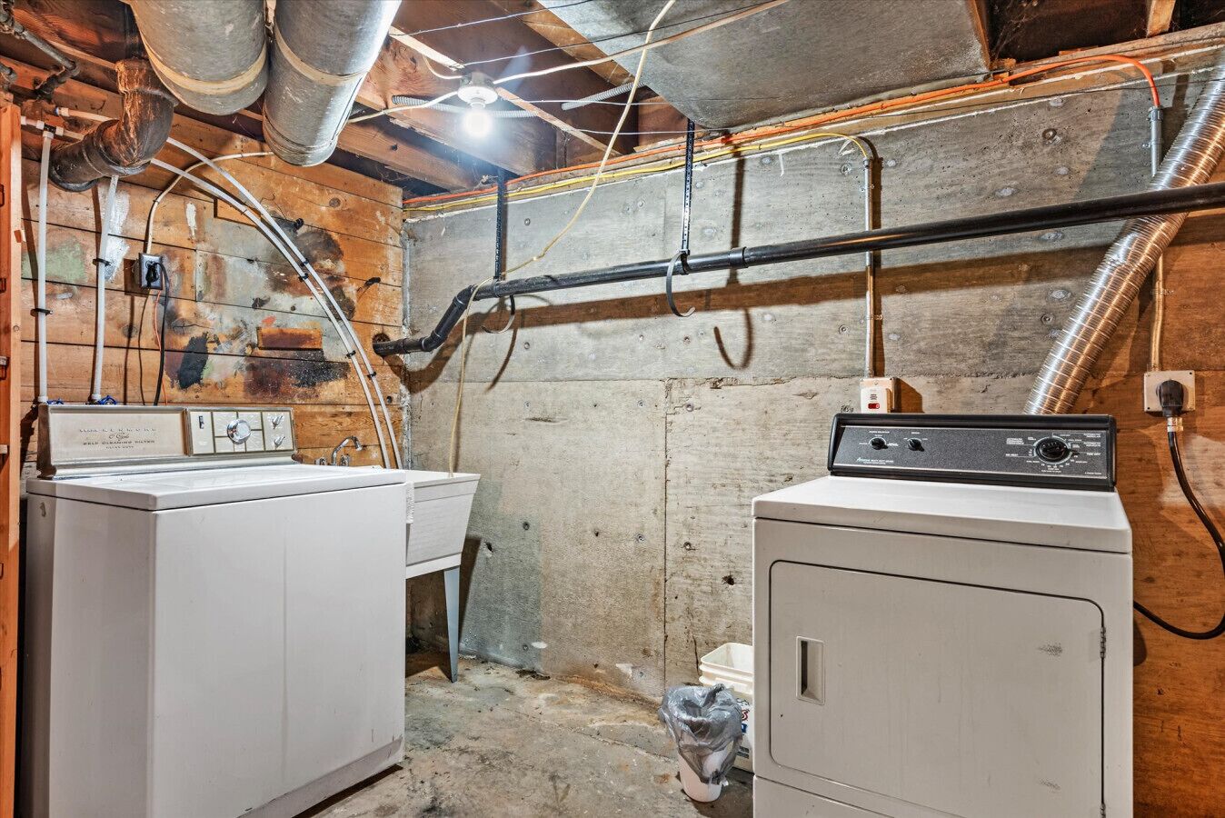A laundry room with two washing machines, visible pipes, and a concrete floor.