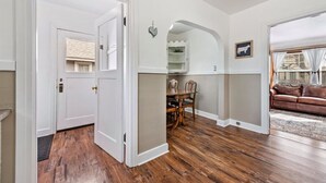 Puerta abierta que da a un comedor con una mesa y sillas de madera, una puerta blanca con un ventanal pequeño y un sofá de cuero marrón en una sala de estar.