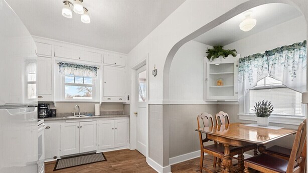Una cocina con armarios blancos, una mesa de comedor de madera y sillas. Hay una ventana con vista a árboles, y una planta en el alféizar.