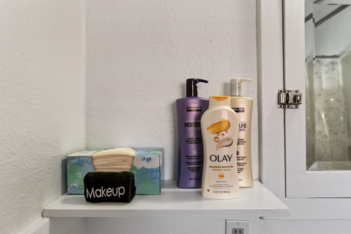 A bathroom shelf with a makeup brush, a box of makeup, and three Kirkland body care products.