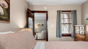 Un dormitorio con cama, cómoda, ventana con cortinas, un cuadro en la pared y una puerta que da a otra habitación.