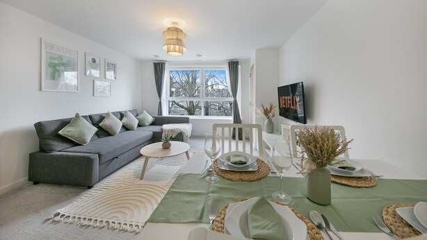 Un salon moderne avec un canapé gris, une table à manger dressée pour deux personnes, un téléviseur fixé au mur et une grande fenêtre donnant sur un paysage arboré.