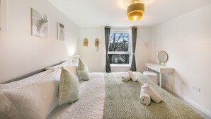 Une chambre à coucher comprenant un lit, deux tables de chevet, une fenêtre avec des rideaux, un luminaire fixé au mur et un miroir.