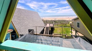 Blick aus einem Fenster auf einen Strand, eine Holzterrasse und ein Haus mit geneigtem Dach.