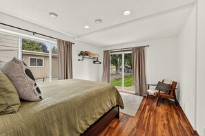 A bedroom with a large bed, a chair, a window with curtains, and a view of the outdoors.