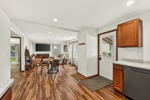 A modern kitchen with wooden cabinets, a dining area with a round table and chairs, and a hallway leading to a living space.