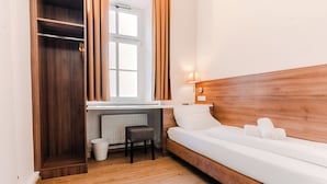 Un dormitorio con cabecera de madera, cama blanca, escritorio con silla, radiador y ventana con cortinas.