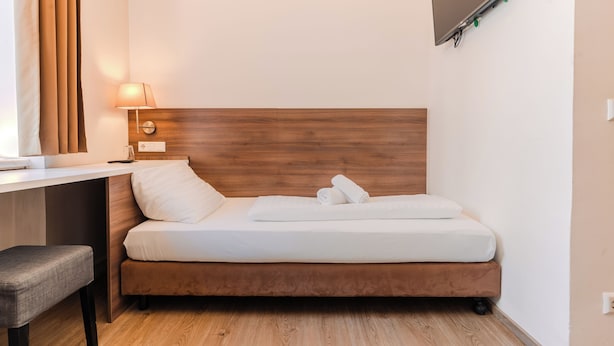 Habitación de hotel pequeña con cama de cabecera de madera, escritorio con lámpara y televisor de pantalla plana montado en la pared.