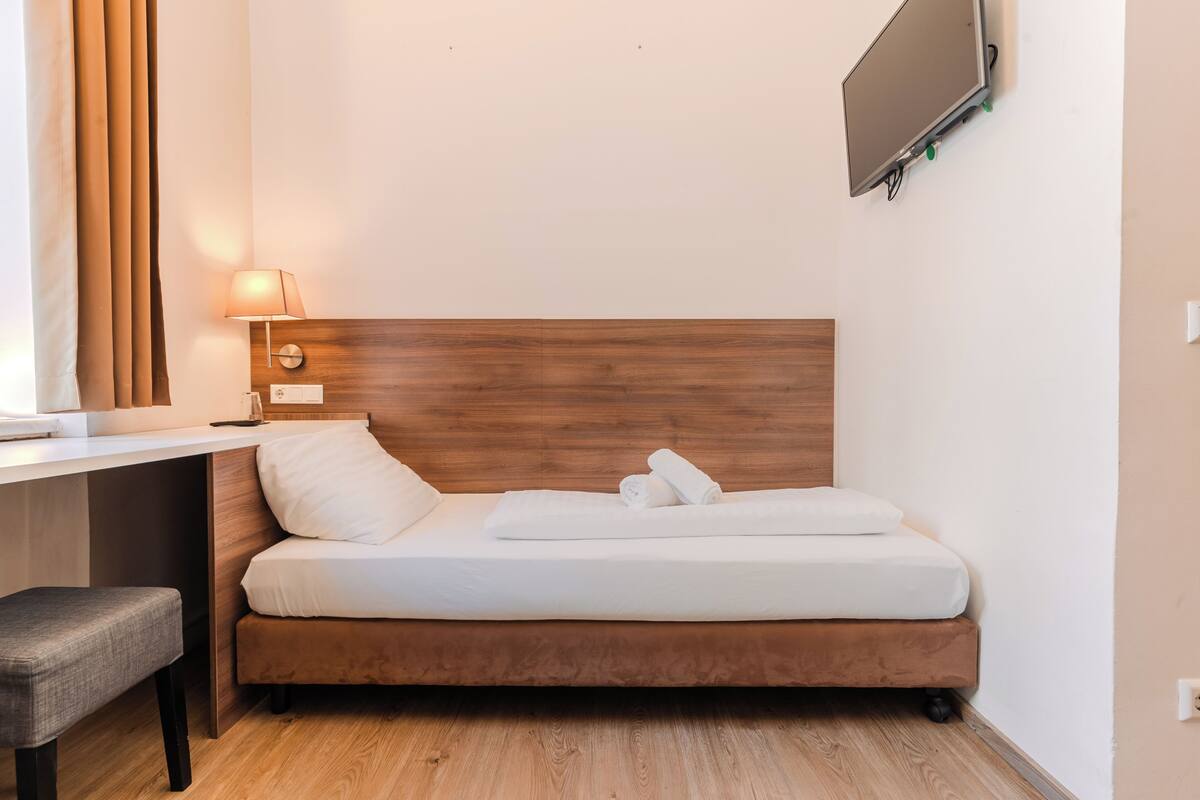 A compact hotel room with a wooden headboard bed, a desk with a lamp, and a flat-screen TV mounted on the wall.