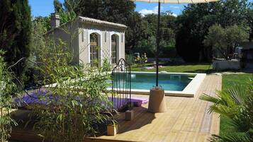 Un espace piscine avec une terrasse en bois, un petit bâtiment doté de fenêtres en arc de cercle et un grand parasol.
