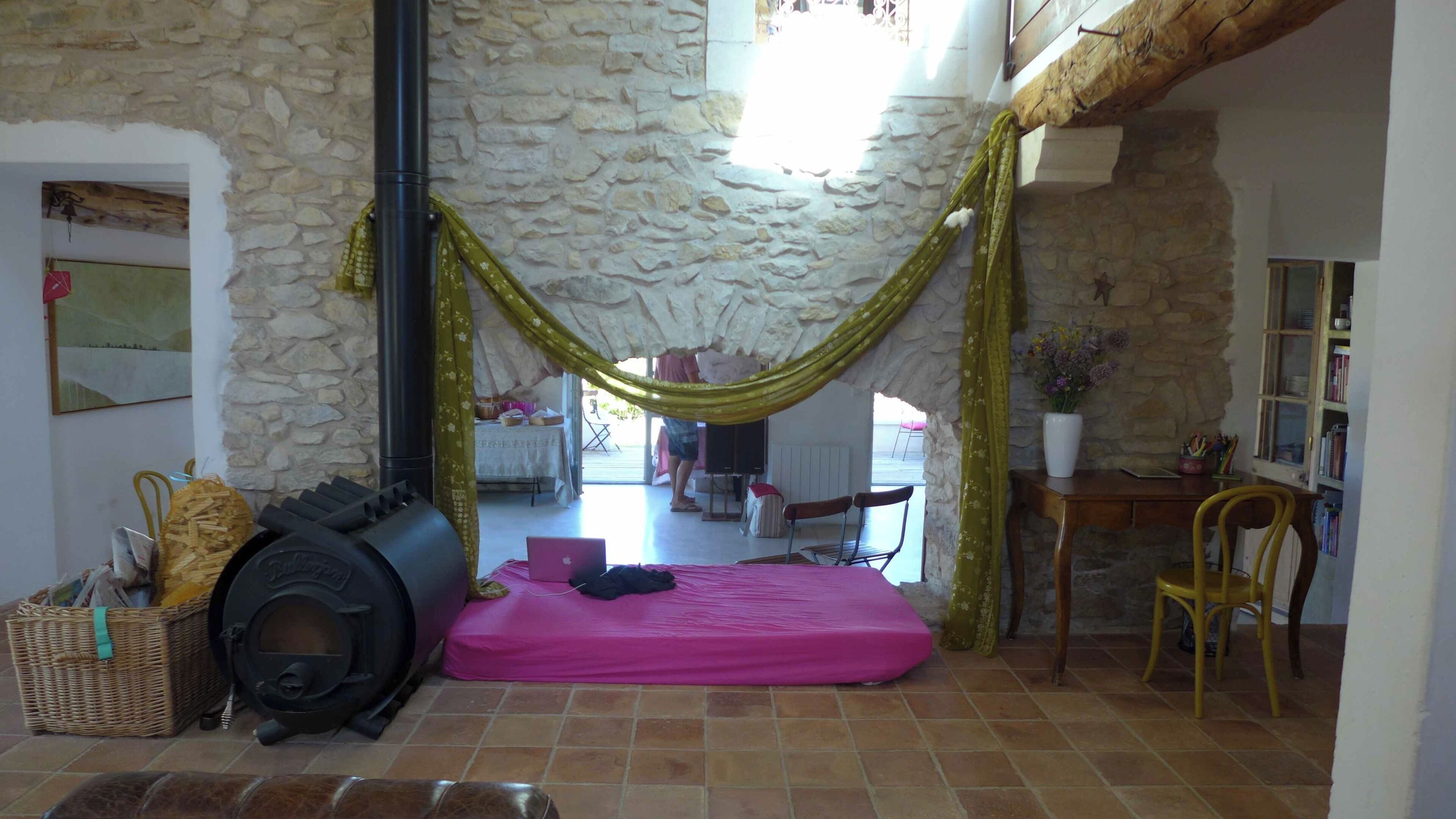 Une pièce avec un mur en pierre, un hamac, un lit, une cheminée et un bureau avec une chaise.