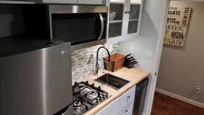 Una cocina con refrigerador de acero inoxidable, microondas, estufa a gas y un lavamanos. Hay armarios sobre la estufa y una encimera con un bloque para cuchillos.