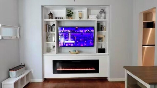 Una sala de estar con un televisor de pantalla plana sobre un mueble blanco, una chimenea abajo y piso de madera.