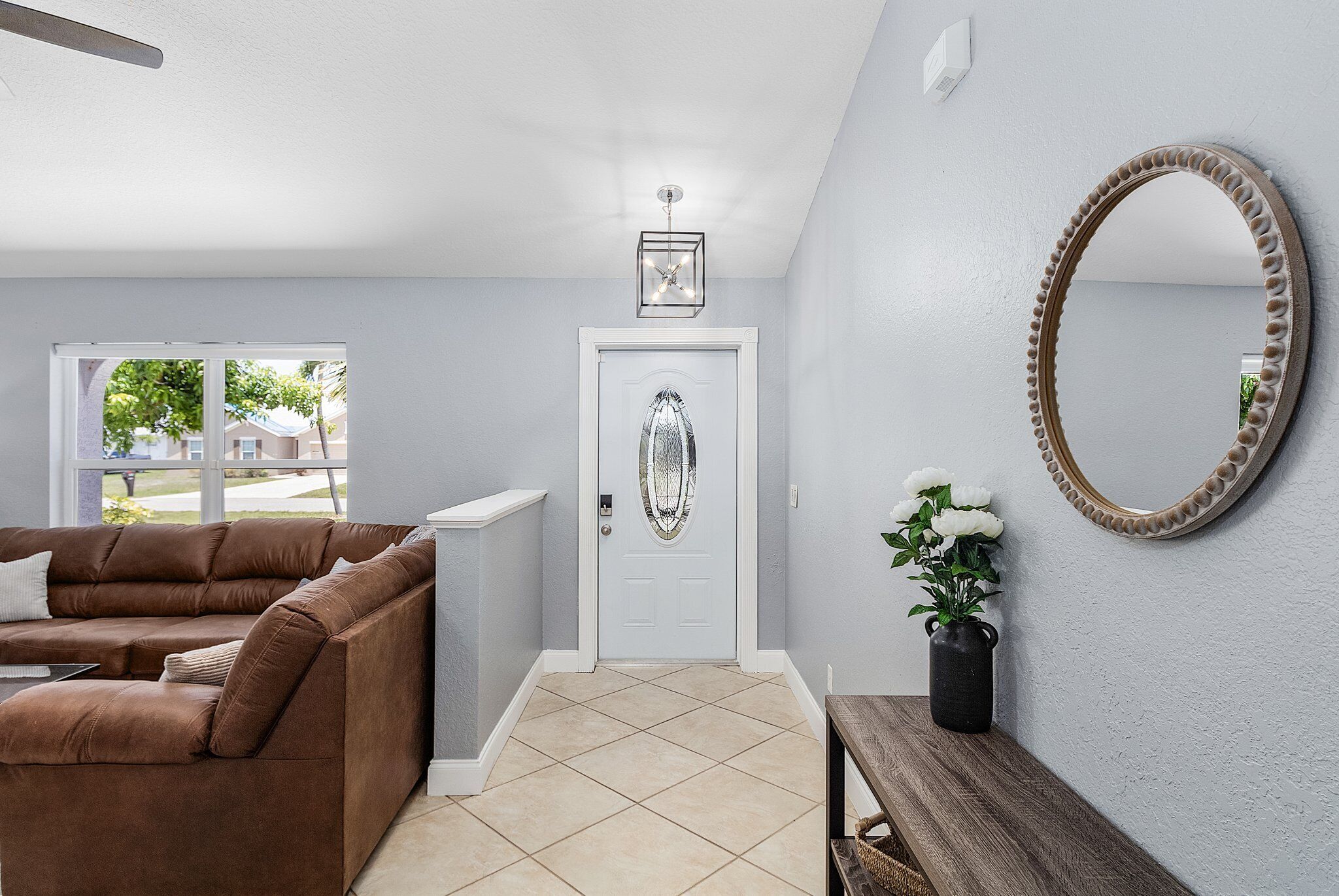 Un couloir avec un canapé marron, une porte blanche, un miroir et un vase rempli de fleurs.