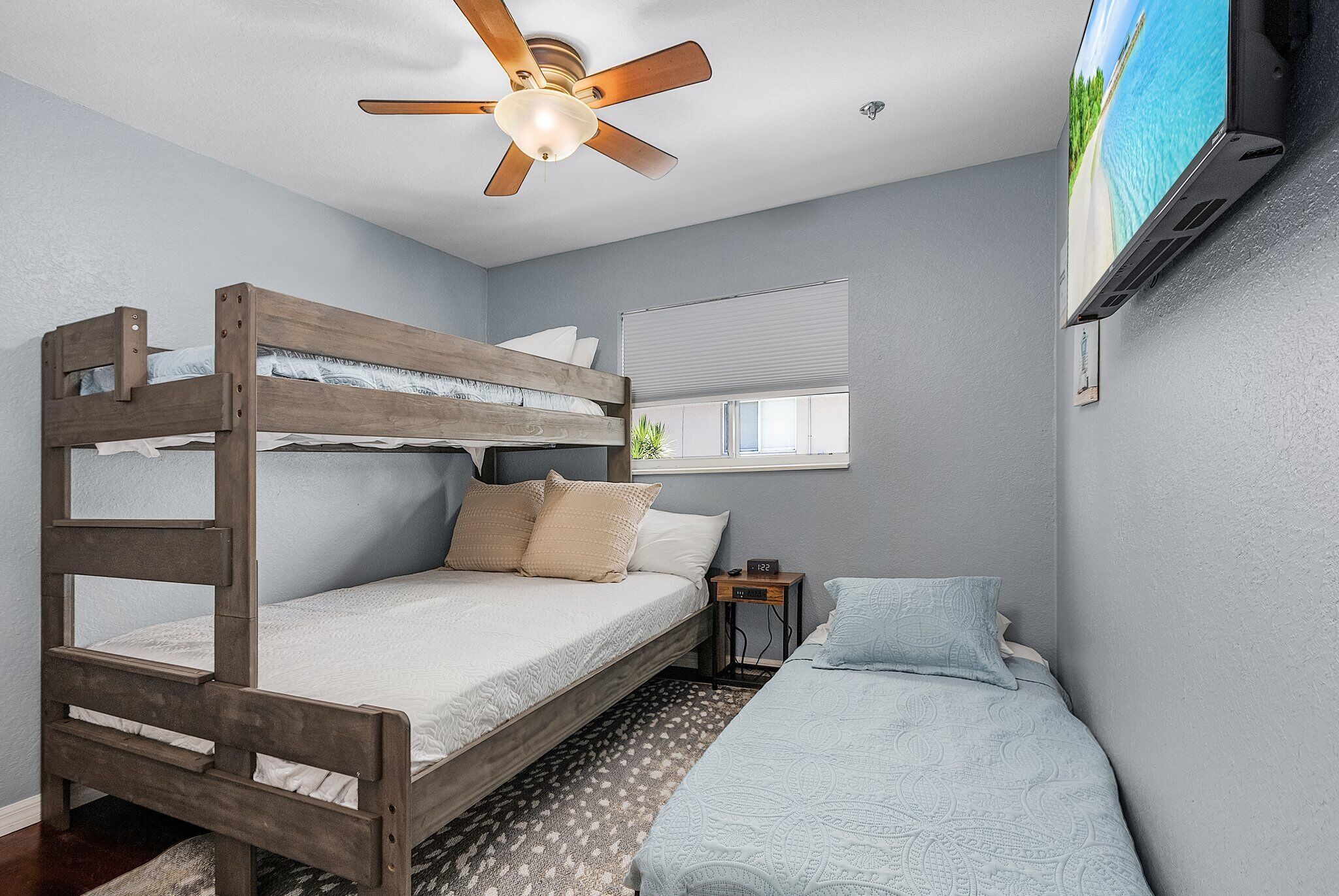 A bunk bed room with a ceiling fan, a TV mounted on the wall, and a window with blinds.