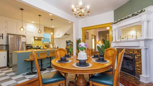 A dining area with a round wooden table, four chairs, a chandelier, a fireplace, and a kitchen in the background. - Near Starland, River St & DT - Gaming Arcade - Large Yard (Savannah)