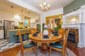 Ein Essbereich mit einem runden Holztisch, vier Stühlen, einer Chandelüster, einem Kamin und einer Küche im Hintergrund.