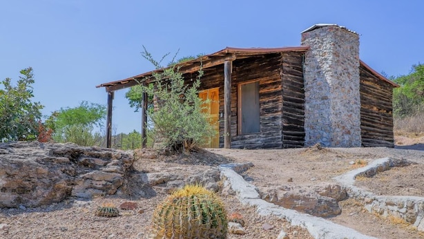A rustic log cabin with a stone chimney, surrounded by desert vegetation and rocky terrain. - Centro de Aventuras Tatu (San Miguel de Allende)
