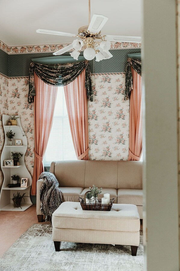 Une pièce avec un canapé beige, un pouf blanc, un ventilateur de plafond et un papier peint à motifs floraux.