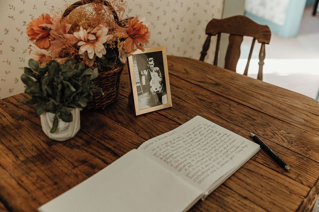 A wooden table with a book, a pen, a framed photo, and a vase with flowers.