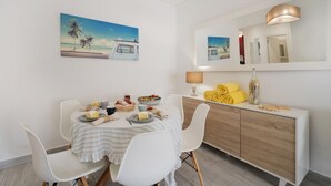 A dining area with a table set for two, a white cabinet with yellow towels, and a painting of a beach scene.