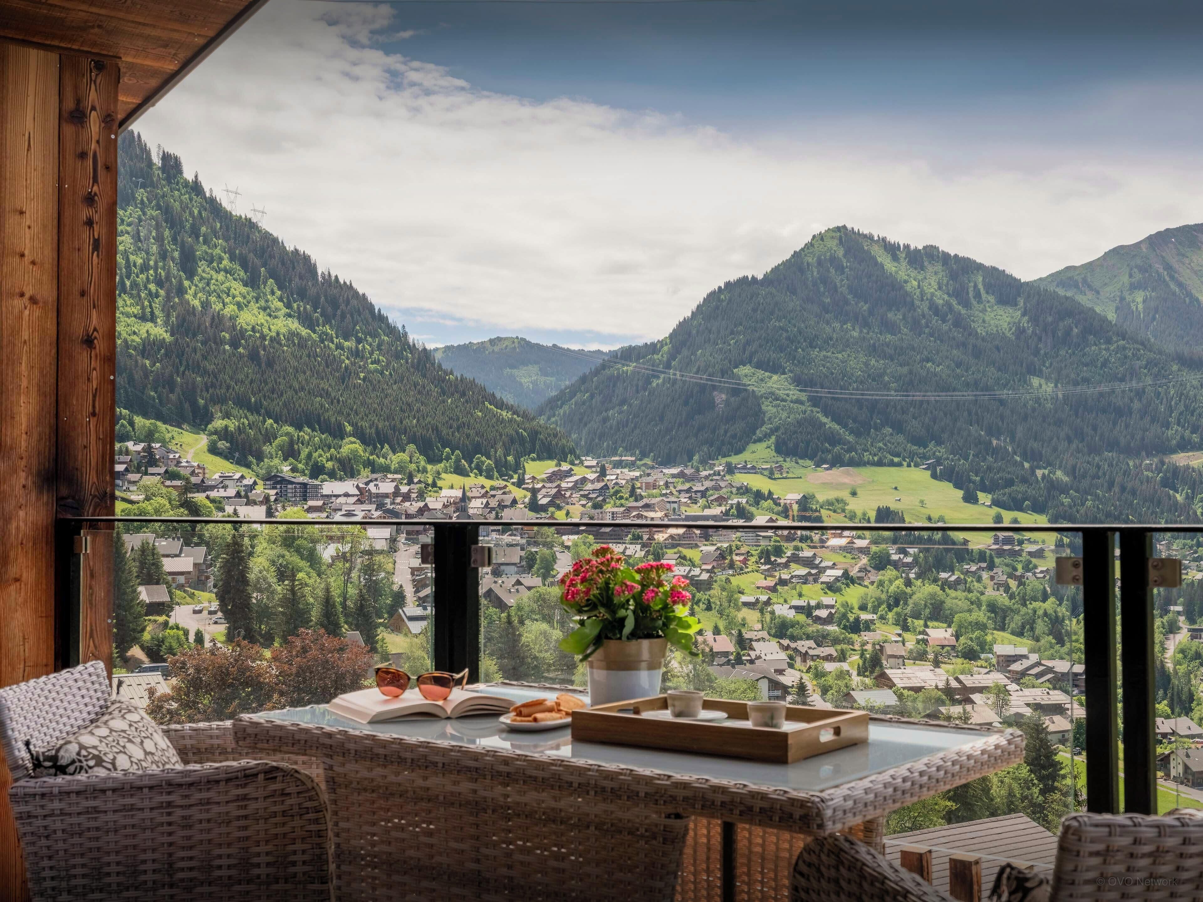 Een balkon met rieten meubels, een tafel met een boek, glazen en een dienblad, en uitzicht op een stad omringd door bergen.