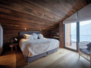 Une chambre avec un plafond en bois, un lit recouvert de draps blancs, un petit bureau avec un ordinateur, et une vue sur un paysage enneigé par une grande fenêtre.