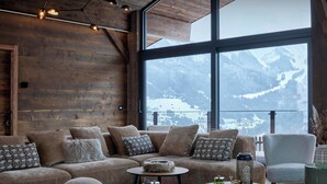 A cozy room with a wooden wall, a sofa set, two round tables, and a view of snow-covered mountains.