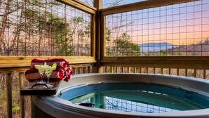 A hot tub with a wooden deck, a table with drinks, and a view of trees and mountains.