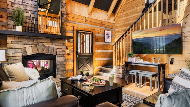 A cozy log cabin interior with a stone fireplace, wooden walls, a dining area, and a large window with a painting.