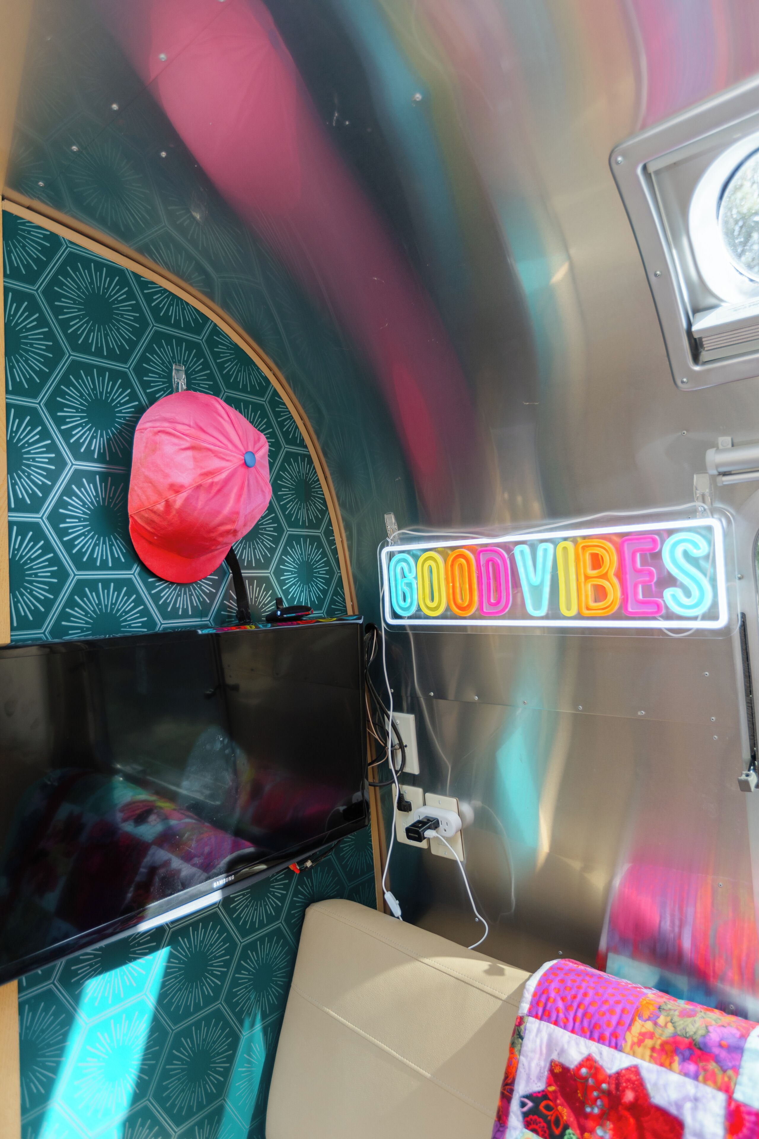 A room with a neon sign reading 'GOODVIBES,' a pink cap hanging on the wall, and a patterned headboard.