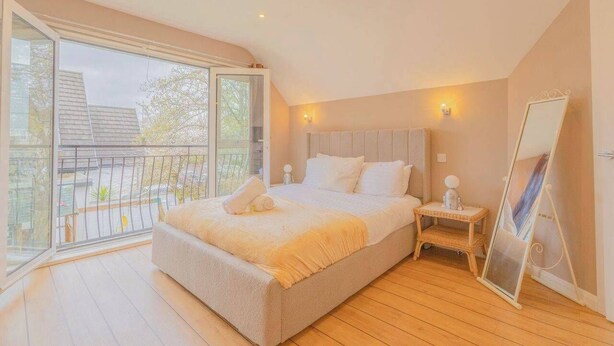 A bedroom with a large bed, a bedside table, a mirror, and a balcony with a view of buildings and trees. - 6BR Mansion With Games Room & Hot Tub (Cardiff)