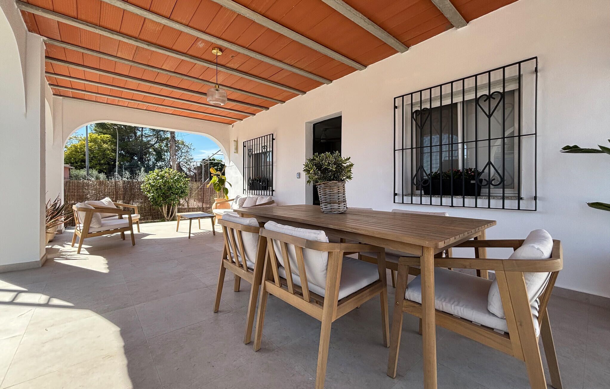 Eine überdachte Terrasse mit einem Holztisch und Stühlen, einem Blumentopf und Blick ins Freie.
