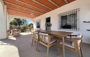 Un porche techado con una mesa y sillas de madera, una planta en maceta y vistas al exterior.