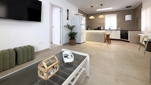 Ein modernes Wohnzimmer mit Flachbildfernseher, Glastisch, grünem Sofa und Küchenbereich mit Kühlschrank, Mikrowelle und Herd.