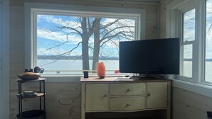 A room with a wooden desk, a television, and a view of a lake and trees through a large window.