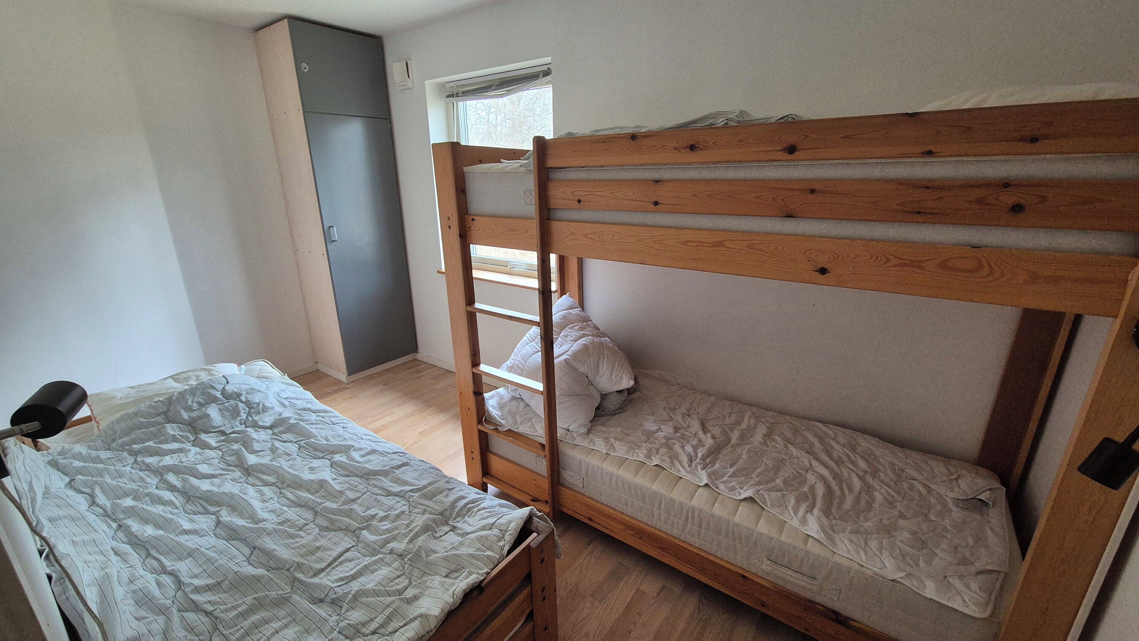 Una cama litera con estructura de madera, una cama individual con una manta a rayas, una escalera de madera, un armario y una ventana con persianas.