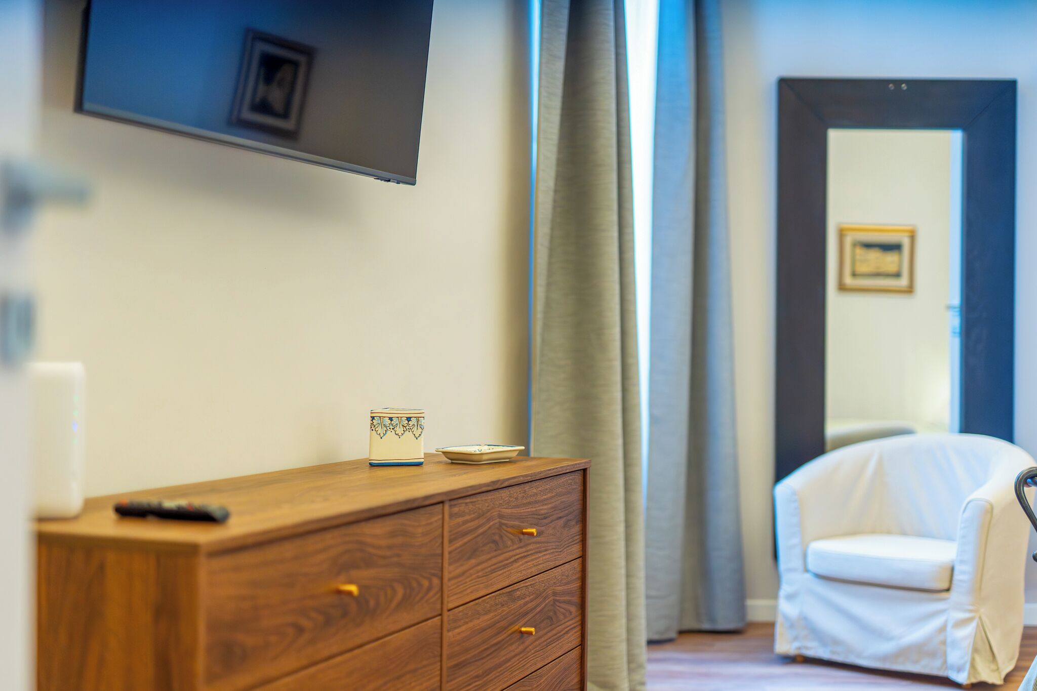 A hotel room with a wooden dresser, a television mounted on the wall, a white chair, and a door with a framed picture.