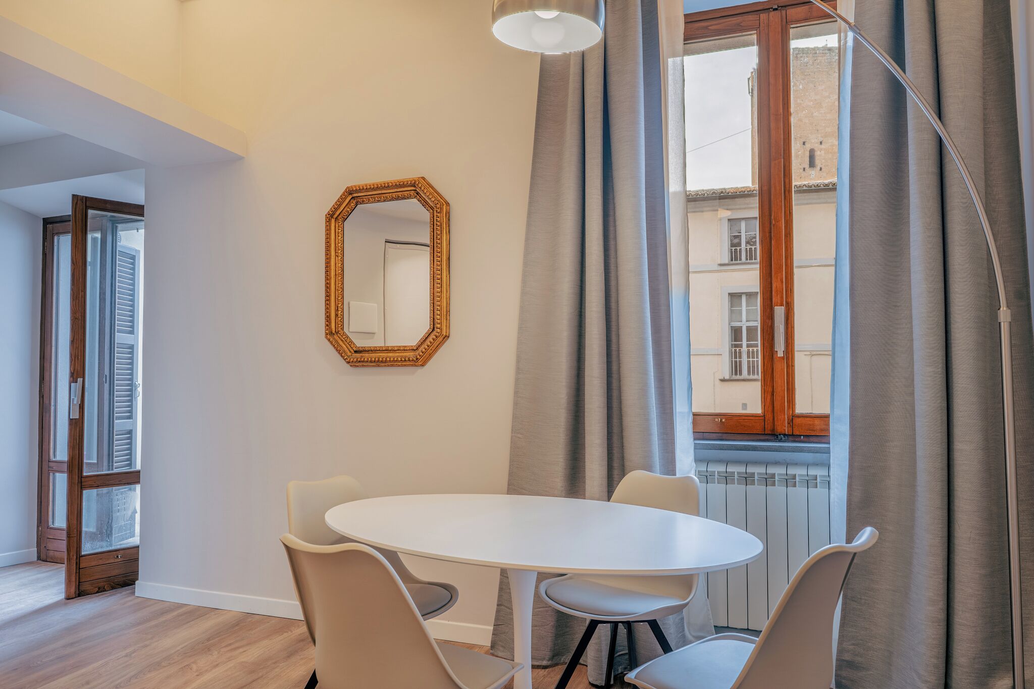 A room with a round table, four chairs, a mirror, and a window with curtains.