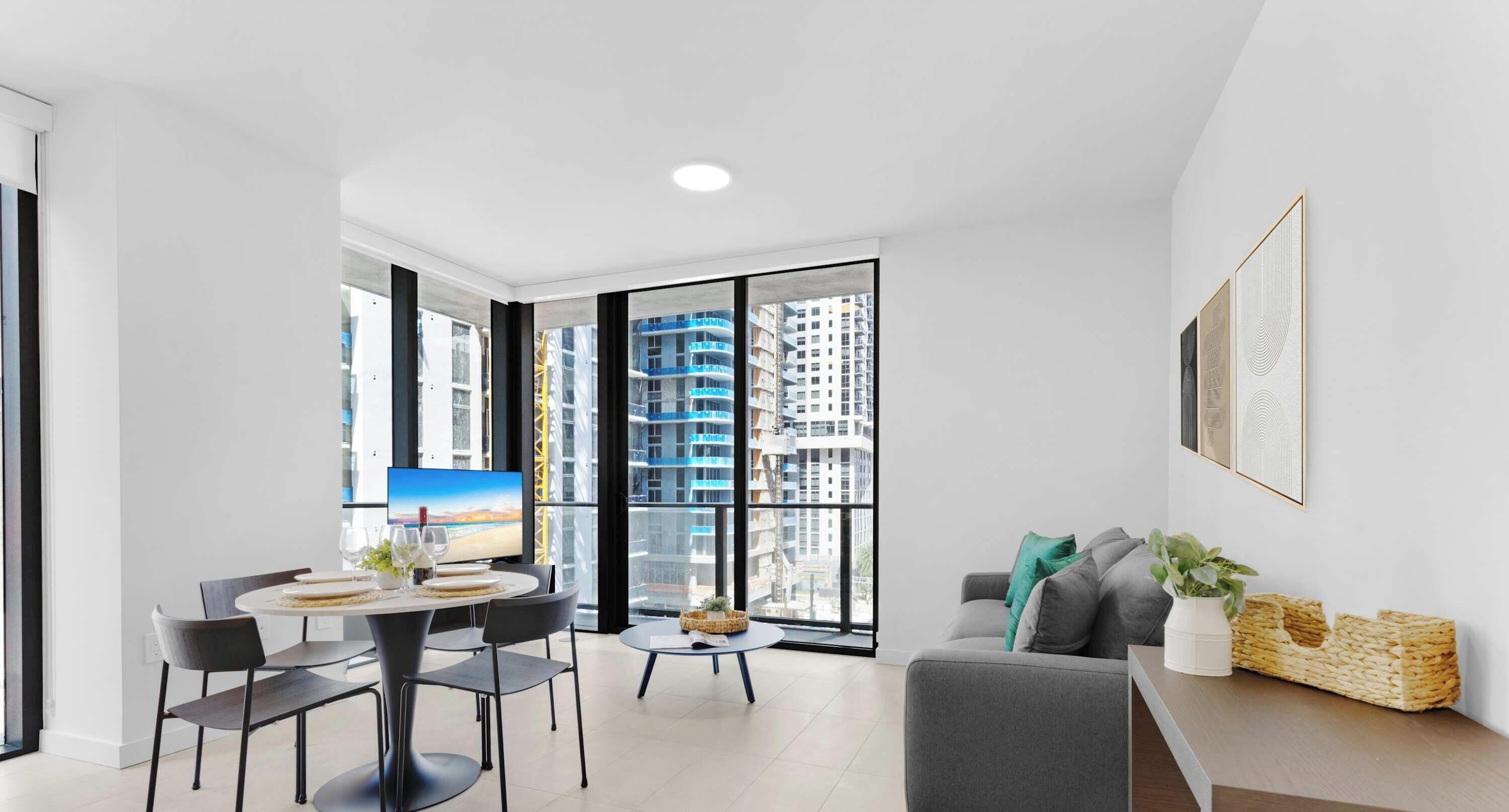A modern living room with a dining area, large windows, a flat-screen TV, and a sofa with cushions.
