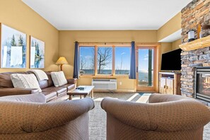 Un salon comprenant un canapé, des fauteuils, une cheminée, un téléviseur et une grande fenêtre donnant sur un beau paysage.
