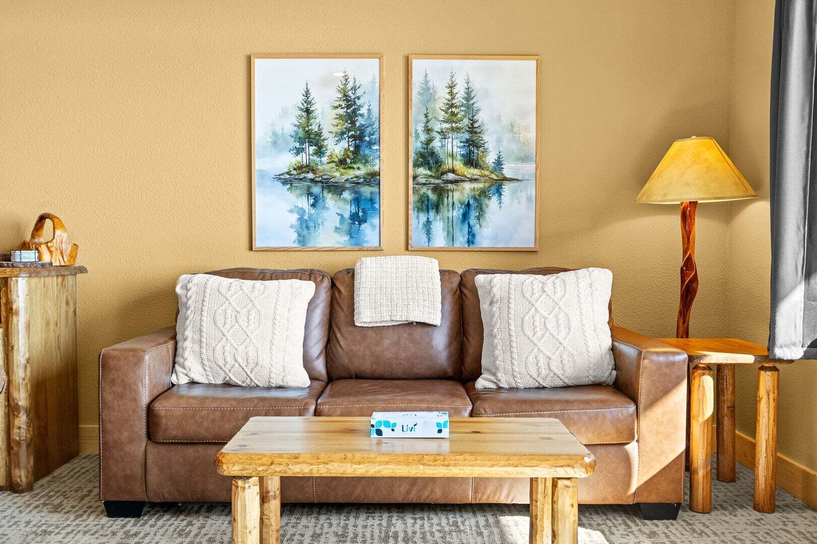 Eine braune Ledersofa mit drei Kissen, ein Holztisch, zwei gerahmte Gemälde von Bäumen und Wasser, eine Lampe und ein Holzhochentisch.