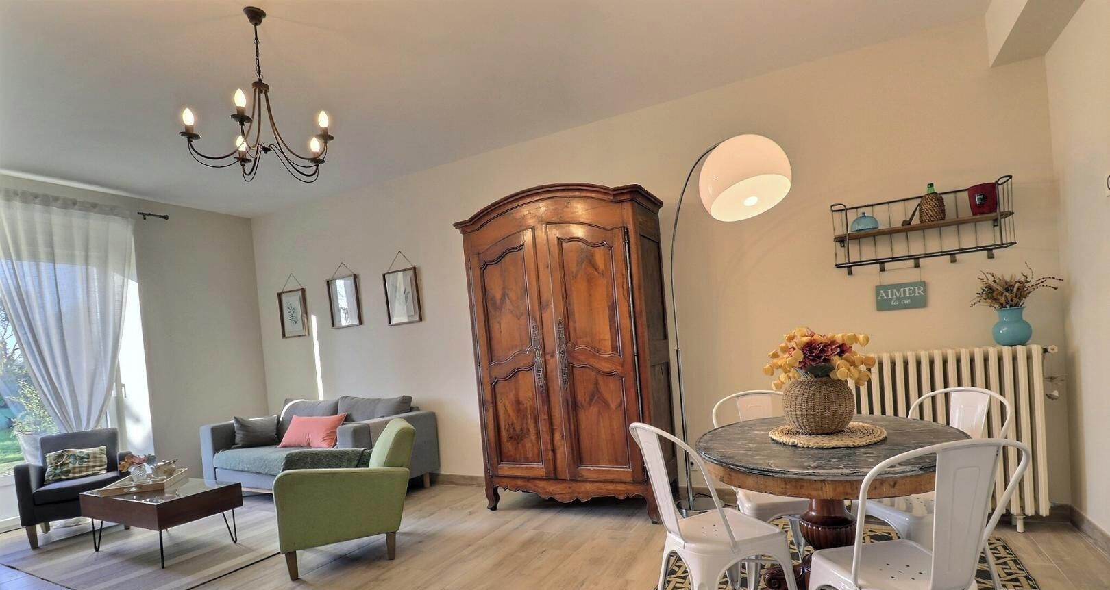 A living room with a sofa, armchair, coffee table, dining table with chairs, a wooden cabinet, a chandelier, a floor lamp, and a radiator.