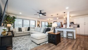 A modern living room with a sectional sofa, a ceiling fan, and a kitchen area with white cabinets and a bar. - 5BR/3BA Stylish Modern Home in Decatur (Decatur)