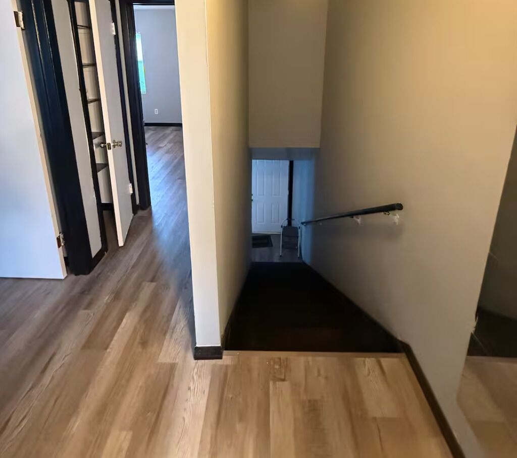 Un couloir avec un plancher en bois, une main courante noire et une porte blanche sur le côté gauche.