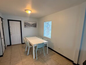A room with a white table and chairs, a door, a window with blinds, and a ceiling light.