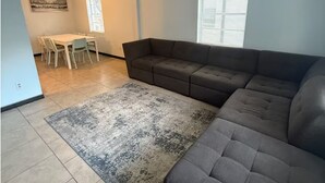 Una sala de estar con un sofá modular, una mesa de comedor con sillas, una alfombra y un cuadro en la pared.