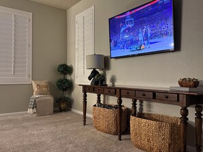 Un salon avec un téléviseur fixé au mur diffusant un match de basket-ball, une console en bois sur laquelle est posée une lampe, et des paniers en osier.