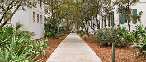 A wooden pathway flanked by greenery and white buildings with green shutters.